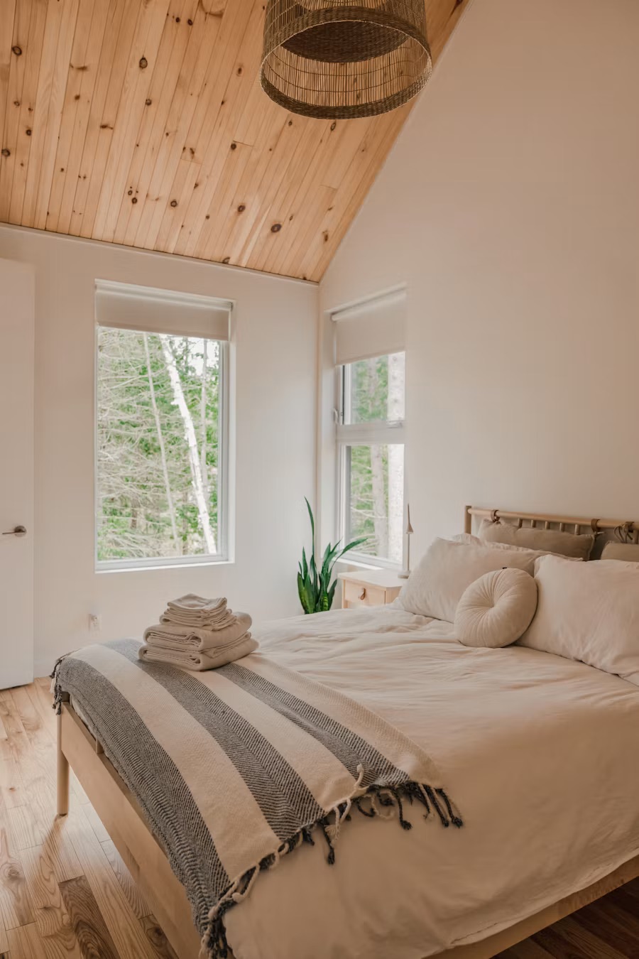 Bright bedroom with a wood-paneled vaulted ceiling, made bed with folded towels, and windows onto trees